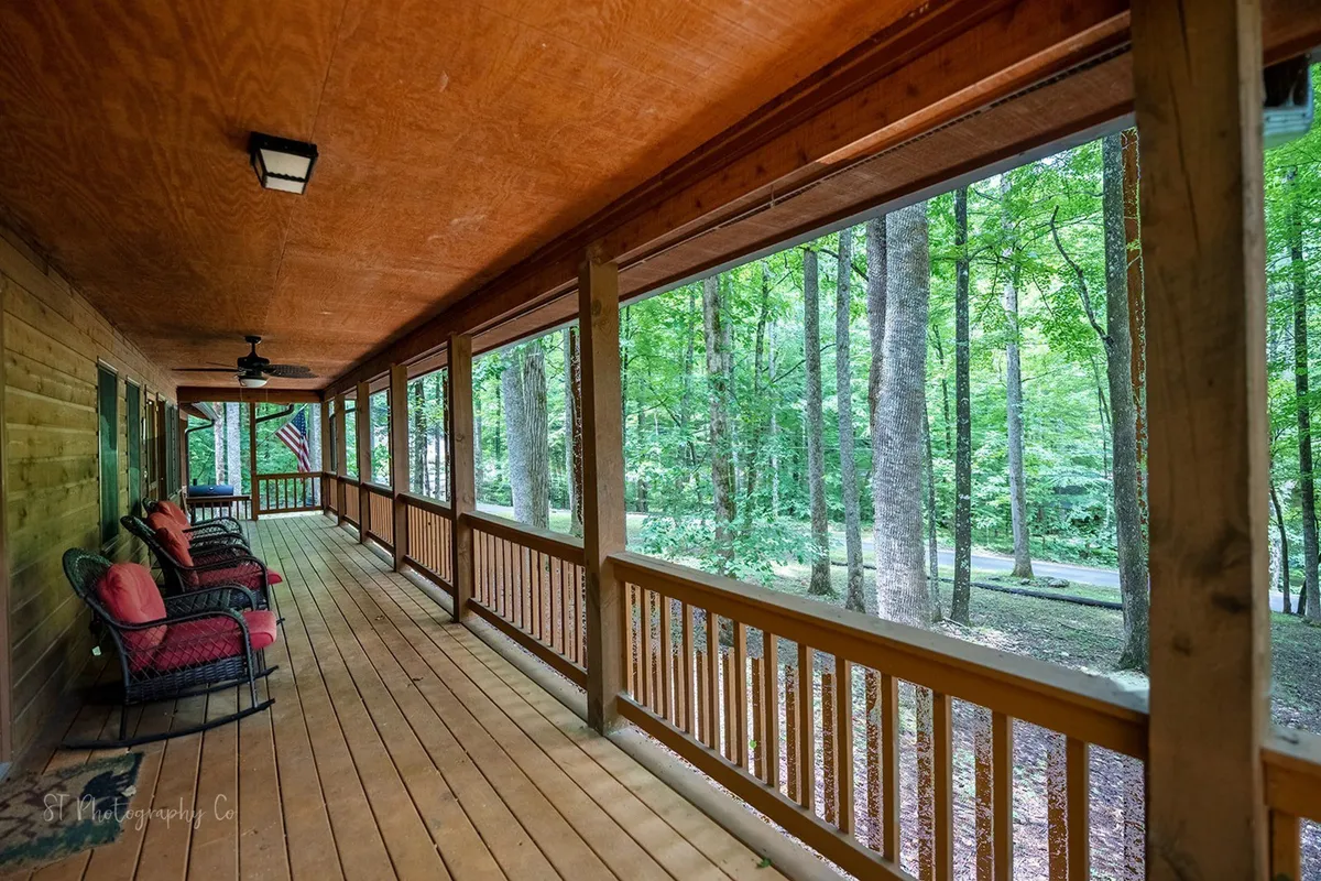 This exterior shot focuses on a pair of cushioned rocking chairs positioned on a wooden deck, bordered by classic railings and surrounded by dense, mature trees. The inviting set-up suggests a perfect spot for morning coffee or quiet reflection while immersed in natural beauty.