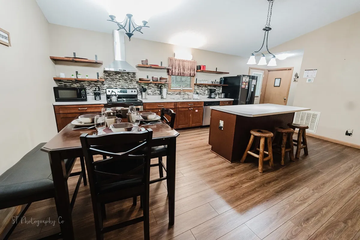 A dining area set for a family meal, showcasing a wooden dining table with matching chairs, a kitchen island with stools, modern appliances, and rustic decor accents. The space feels warm, inviting, and ideal for sharing meals and stories.