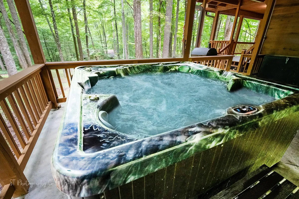 This exterior image showcases a hot tub set on a spacious wooden deck, surrounded by lush Smoky Mountain trees. The tranquil setting invites relaxation and offers a panoramic view of the natural landscape, seamlessly blending indoor comfort with outdoor living.
