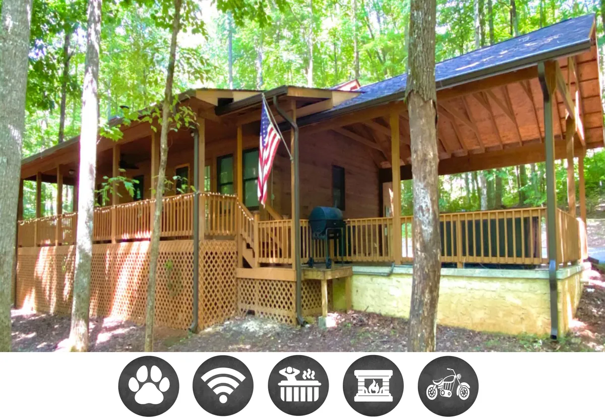 A charming wooden cabin nestled among towering trees, featuring a spacious deck with a grill, an American flag fluttering in the breeze, and serene woodland views—perfect for outdoor relaxation and barbecues after a day in the Smoky Mountains.