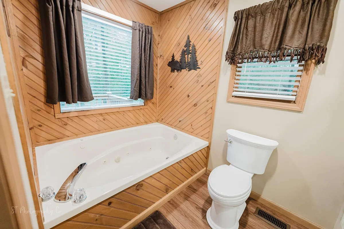 A tranquil bathroom scene with a whirlpool jetted tub surrounded by wood paneling, a window with curtains offering a view outside, and decorative wall art. The setting suggests relaxation and a soothing soak after a long day.