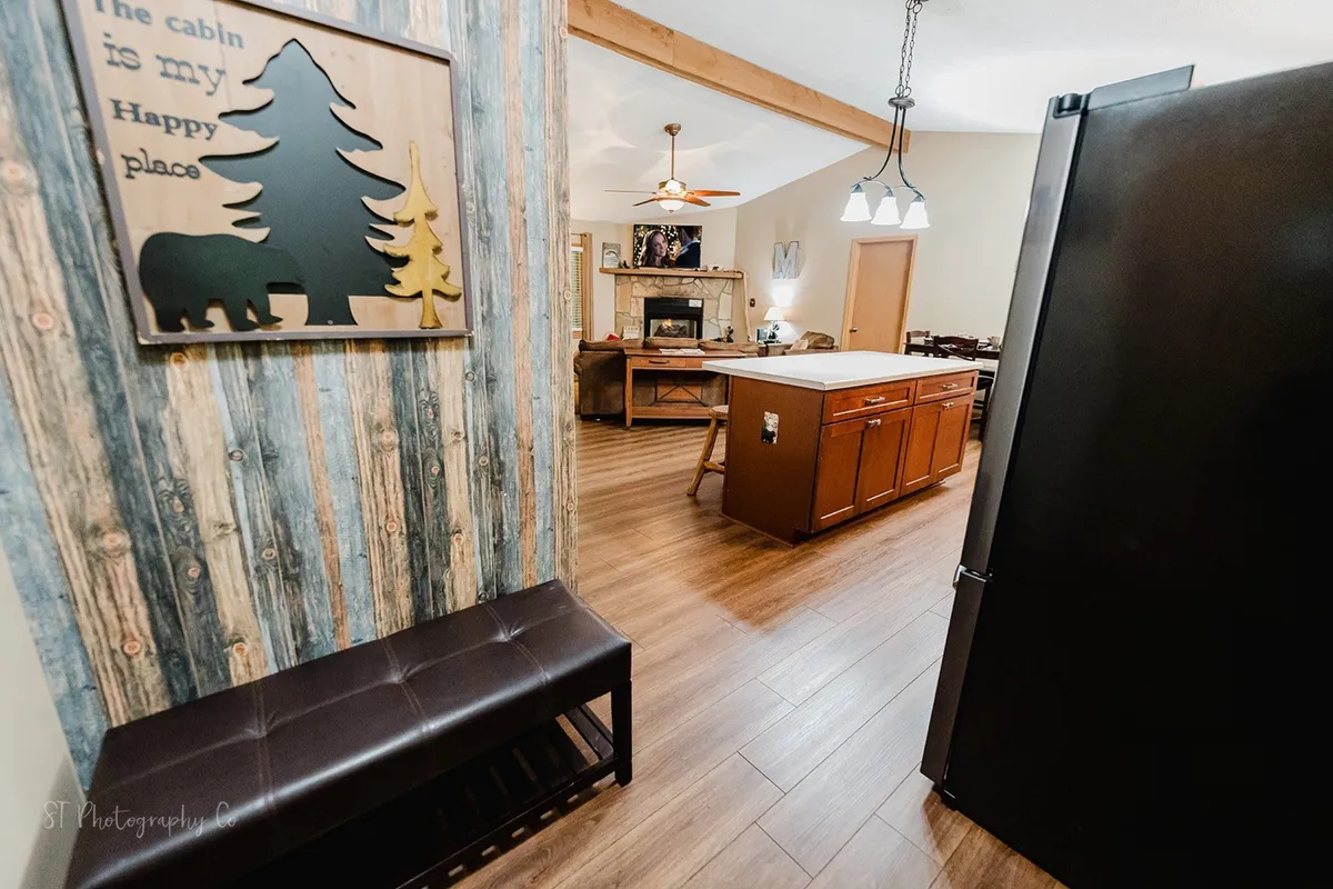 A warm and welcoming open-plan area featuring a kitchen island, brown leather bench, and wooden wall art with trees and a bear. The space is designed for family gatherings, with inviting spots for relaxing by the fireplace or enjoying cozy meals together.