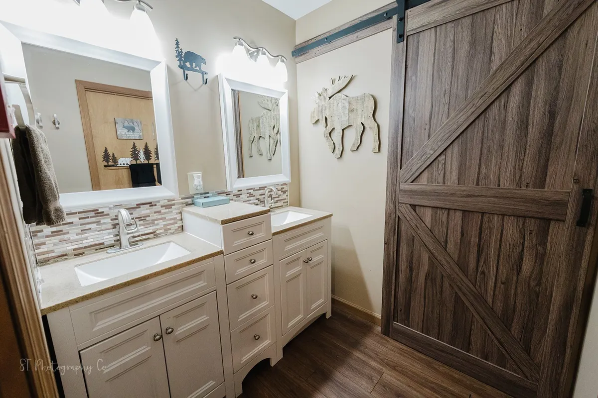 A bright bathroom featuring a double sink vanity with mirrors, barn door, and playful moose wall decor. The ambiance is fresh and inviting, ideal for unwinding or morning routines.