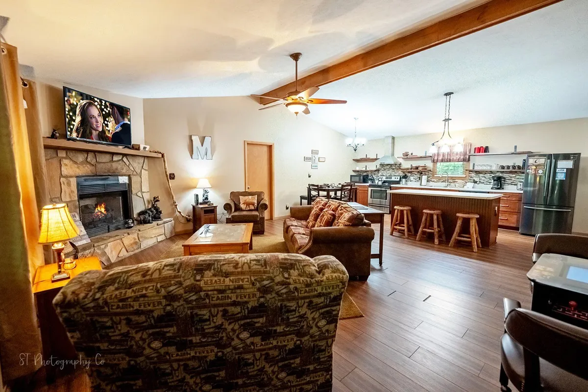 A spacious open-concept living area featuring a cozy stone gas fireplace, comfortable sofa set, flat-screen TV, and a kitchen island that seamlessly connects to the kitchen and dining spaces. The room is warmly lit, with rustic accents and inviting seating arranged for conversation, relaxation, or movie nights.