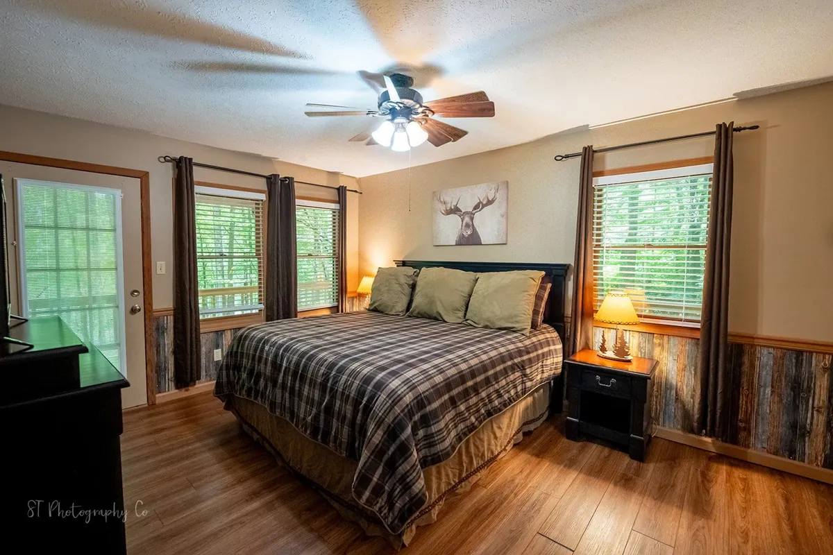 A cozy bedroom with plaid bedding, wooden furniture, and a window offering natural light and views of the outdoors. The decor includes a decorative moose artwork, adding to the mountain retreat atmosphere.