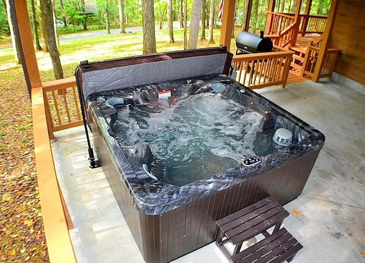 A view of a wooden deck featuring a hot tub, ample space for gathering, and towering trees encircling the area. The setup suggests opportunities for both relaxation and social activities, perfectly capturing the spirit of outdoor living in a private, wooded setting.
