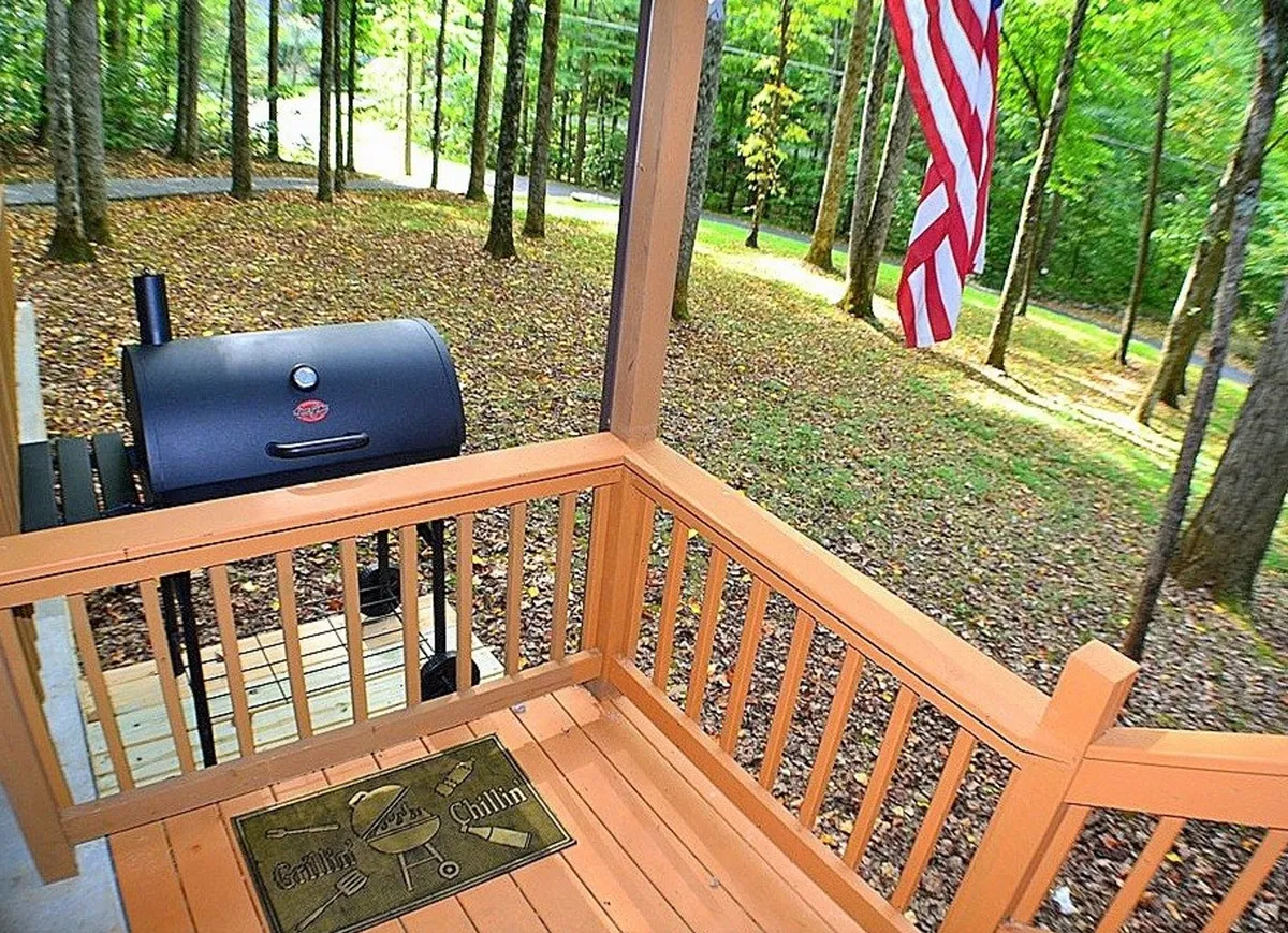 This exterior image highlights a charcoal grill set up on the porch, complemented by a welcoming mat and flag, with tall trees providing a natural backdrop. The setting is primed for evening barbecues or casual gatherings, linking hospitality with outdoor enjoyment.