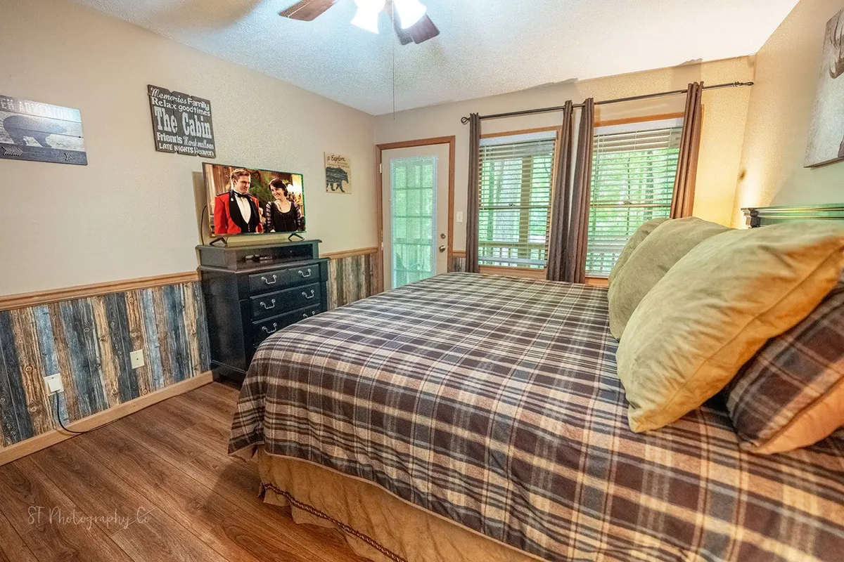 A warmly lit bedroom featuring a bed with a plaid bedspread, wooden furniture, and large windows with blinds. There's a television showing a movie, and the room is filled with natural light, offering a cozy and tranquil retreat.