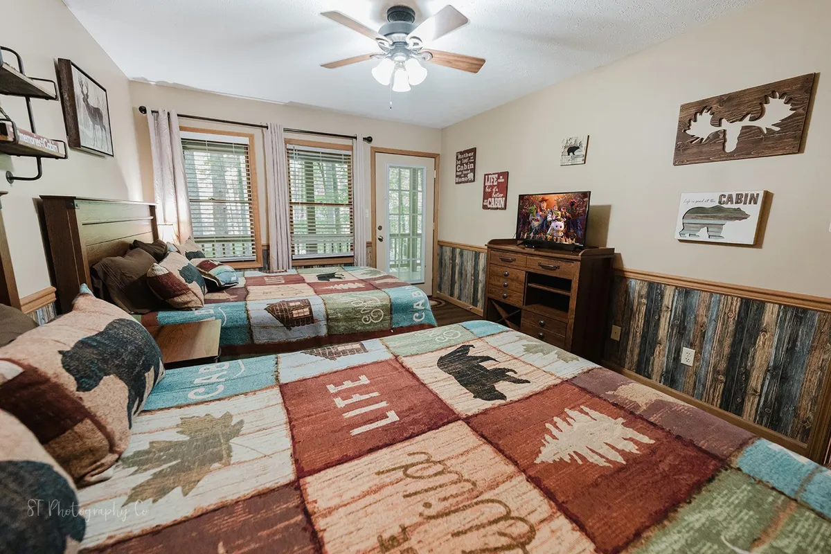 A spacious bedroom with two double beds adorned with patterned quilts, a wooden dresser with a TV, and large windows allowing in plenty of light. The mood is rustic and comfortable, ideal for families or groups.